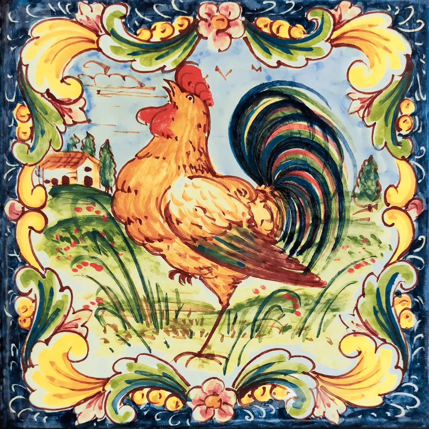Azulejo decorado con un gallo en un paisaje rural, rodeado de un marco azul con hojas amarillas y verdes y pequeñas flores rosas